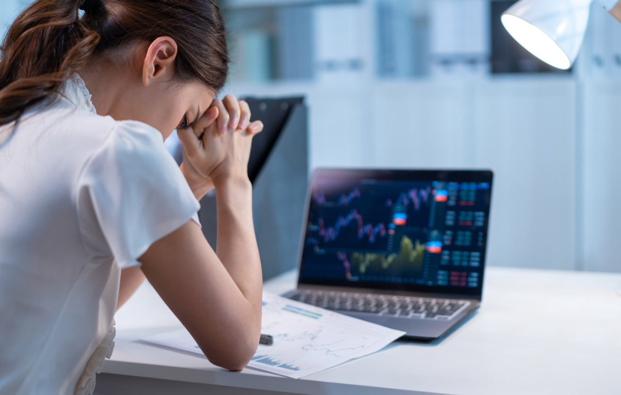 How to Overcome the Fear of Losing Your Gains — a woman sitting at a desk with her hands clasped and head bowed in frustration.